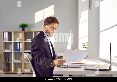 Fokussierter, seriöser Geschäftsmann, der am Laptop sitzt und sich Papierdokumente ansieht Stockfoto