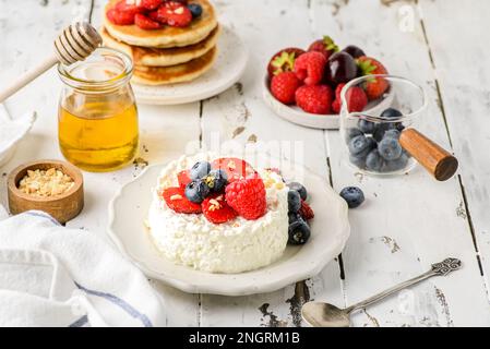 Hüttenkäse mit Himbeeren, Erdbeeren und Heidelbeeren. Hüttenkäse wird in Form eines Gebäckrings neben einem Glas Honig und Cho aufgemacht Stockfoto