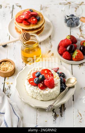 Hüttenkäse mit Himbeeren, Erdbeeren und Heidelbeeren. Hüttenkäse wird in Form eines Gebäckrings neben einem Glas Honig und Cho aufgemacht Stockfoto