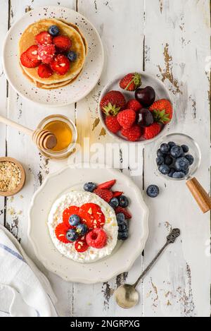 Hüttenkäse mit Himbeeren, Erdbeeren und Heidelbeeren. Hüttenkäse wird in Form eines Gebäckrings neben einem Glas Honig und Cho aufgemacht Stockfoto