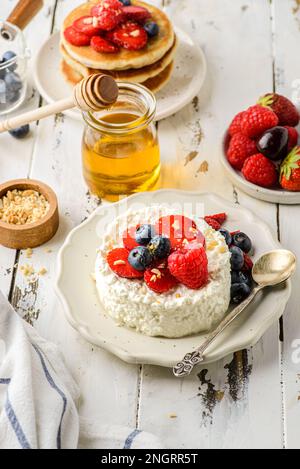 Hüttenkäse mit Himbeeren, Erdbeeren und Heidelbeeren. Hüttenkäse wird in Form eines Gebäckrings neben einem Glas Honig und Cho aufgemacht Stockfoto