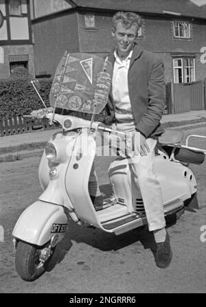 Ein modisch gekleideter junger Mann auf einem Lambretta Roller in Großbritannien Mitte der 1960er Jahre. Er trägt eine lässige Jacke, eine cremefarbene Hose mit Turnups und Wildlederschuhe. Orte, die der Roller besucht hat, werden als Wimpel auf der Windschutzscheibe angezeigt – darunter Paris und Oxford. Dieses klassische Foto aus der Zeit, als Roller zu einer „Mod“-Transportform mit Großbritanniens Jugendkultur wurden. Dieses Bild ist von einem alten Amateur-35mm-Negativ – ein klassisches 1960er-Foto. Stockfoto