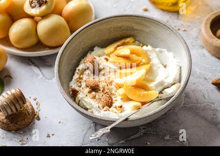 Hüttenkäse mit Aprikosen und Mandeln. Hüttenkäse ist in einer Schüssel auf dem Tisch im Sonnenlicht, neben einem Teller mit Aprikosen und Nüssen und Honig Stockfoto