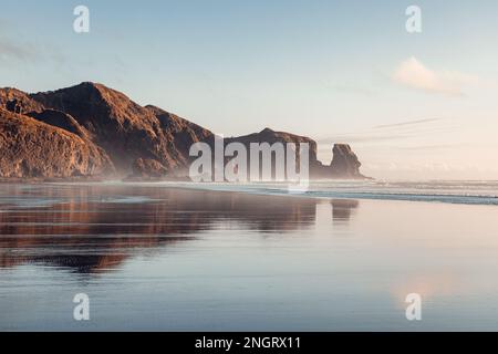 Der Piha Beach ist bei Sonnenuntergang eingefangen, wobei die warmen Farben des Himmels sich auf den ruhigen Wellen des Ozeans spiegeln. Stockfoto