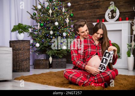 Ein Mann und eine schwangere Frau sehen sich Ultraschallbilder an, während sie auf einem Teppich sitzen. Stockfoto