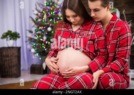 Der Mann, der hinter der Frau sitzt, umarmt sie von hinten und streichelt ihren schwangeren Bauch. Stockfoto