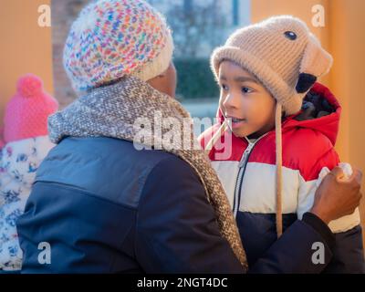 Porträt eines afrikanischen kleinen Kindes in warmer Oberbekleidung farbenfrohes Unterhemd und Strickmütze mit Ohrenklappen und Mutter Pflege und Bindung Stockfoto