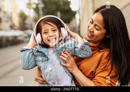 Glückliche filipinöse Mutter mit ihrer Tochter, die im Stadtzentrum mit Kopfhörern Spaß beim Musikhören hat - Familienliebeskonzept Stockfoto