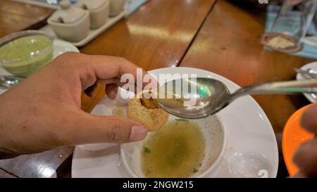 Ein indischer Mann hält Panipuri oder Golgappa oder Puchkas mit Kartoffelmasala und gießt Pani mit silbernem Löffel hinein. Stockfoto