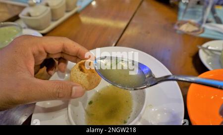 Ein indischer Mann hält Panipuri oder Golgappa oder Puchkas mit Kartoffelmasala und gießt Pani mit silbernem Löffel hinein. Stockfoto