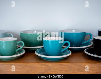 Nahaufnahme Set aus großen und kleinen blau-grünen Tonnen Kaffeetassen, Keramikbecher mit Untertassen stehen in einer Reihe auf einem Holzboden isoliert auf weißem Hintergrund Stockfoto