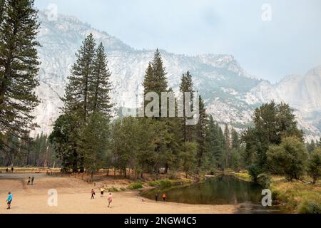 Blick auf den Merced River und die Berge mit Rauch von den Kaminen im Hintergrund. Ein Mann und ein paar Kinder gehen am Fluss entlang Stockfoto