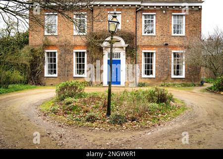 Blick von vorne auf ein altes Herrenhaus mit einer kreisförmigen Auffahrt und einem über schmiedeeisernen Lampenpfosten gebogen inmitten von etwas Grün. Stockfoto