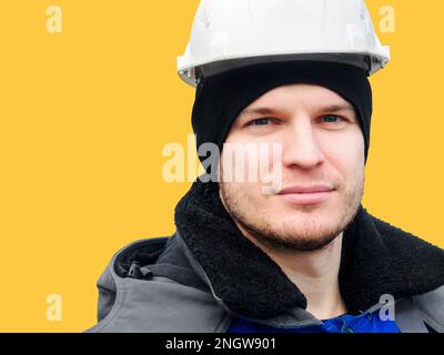 Grosses Porträt des Menschen im Bauhelm auf gelbem Hintergrund. Kaukasischer Mitarbeiter mittleren Alters mit selbstbewusstem Blick blickt direkt in die Kamera. Layout mit Platz zum Kopieren. Stockfoto