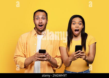 Glückliches, geschocktes junges schwarzes Paar mit offenem Mund, das sich auf Telefonen isoliert auf gelbem Hintergrund unterhält Stockfoto