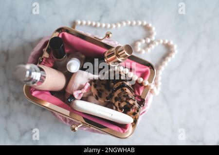 Pinkfarbene Kosmetiktüte, gefüllt mit Make-up-Produkten und Schmuck auf Marmortisch, Nahaufnahme. Schönheitskonzept. Stockfoto