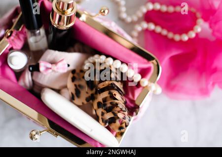 Pinkfarbene Kosmetiktüte, gefüllt mit Make-up-Produkten und Schmuck auf Marmortisch, Nahaufnahme. Schönheitskonzept. Stockfoto