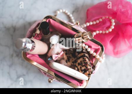 Pinkfarbene Kosmetiktüte, gefüllt mit Make-up-Produkten und Schmuck auf Marmortisch, Nahaufnahme. Schönheitskonzept. Stockfoto