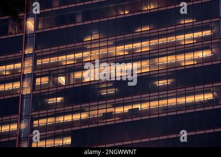 Urbane Geometrie. Nachtsicht auf die verglaste Wand des Hochhauses mit zufällig beleuchteten großen Fenstern und Lampenreihen an der Decke im Gebäude Stockfoto