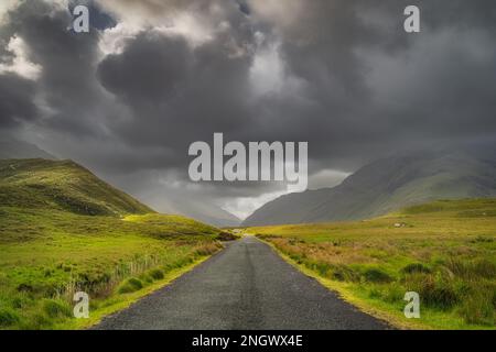 Die Straße führt durch das Doolough Valley, zwischen Glenummera und Glencullin Gebirgsketten, beleuchtet von Sonnenlicht mit dunklem dramatischen Himmel, Mayo, Irland Stockfoto