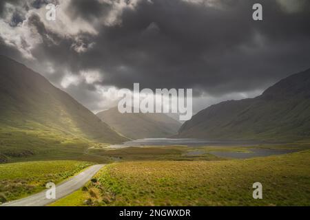 Die Straße führt durch das Doolough Valley mit Seen, Glenummera und Glencullin Bergketten, die von Sonnenlicht und dunklem dramatischen Himmel beleuchtet werden, Mayo, Irland Stockfoto