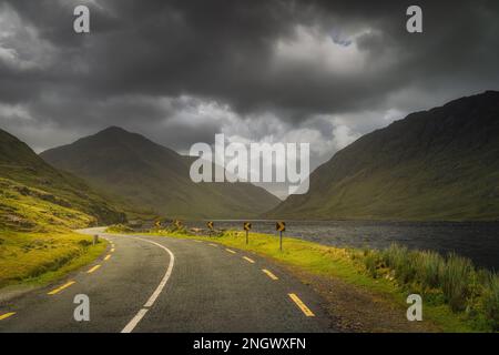 Die Straße führt durch das Doolough Valley neben dem See und zwischen den Bergketten Glenummera und Glencullin mit dunklem dramatischen Himmel, Mayo, Irland Stockfoto