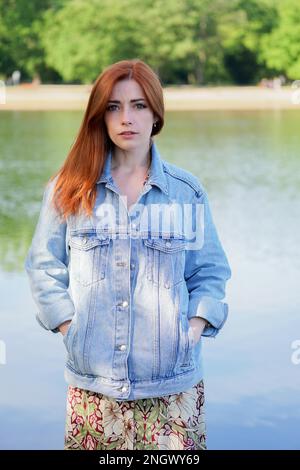 Junge Frau das Tragen von Jeans Jacke über Sommer Kleid stehen am See - authentische Menschen Stockfoto