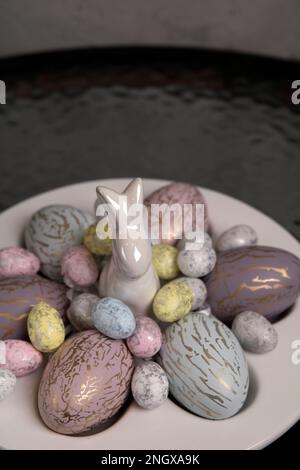 Foto von vielen verschiedenen gefärbten Hühnereiern und wiedergesägten Eiern, die auf einem speziellen Teller für Eier liegen und einem dekorativen Kaninchen, das auf dem Tisch steht Stockfoto