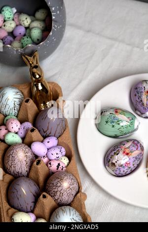 Foto von bunten Eiern unterschiedlicher Größe und einem Kaninchen, das in einem Eierbehälter auf einer weißen Tischdecke steht Stockfoto