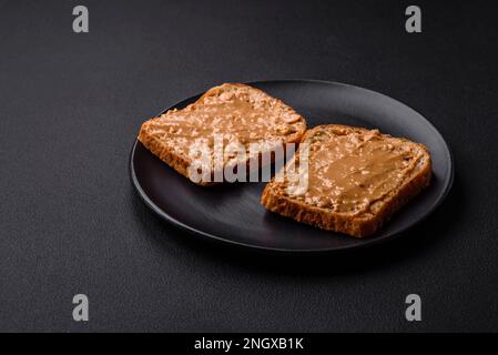 Nahrhaftes Sandwich bestehend aus Brot und Erdnussbutter auf einer schwarzen Keramikplatte auf dunklem Betonhintergrund Stockfoto