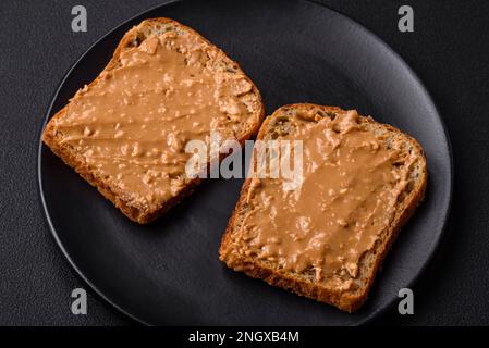 Nahrhaftes Sandwich bestehend aus Brot und Erdnussbutter auf einer schwarzen Keramikplatte auf dunklem Betonhintergrund Stockfoto