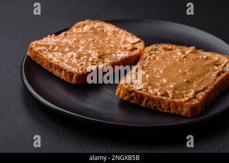 Nahrhaftes Sandwich bestehend aus Brot und Erdnussbutter auf einer schwarzen Keramikplatte auf dunklem Betonhintergrund Stockfoto