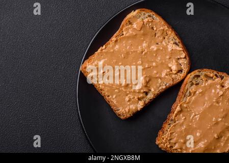 Nahrhaftes Sandwich bestehend aus Brot und Erdnussbutter auf einer schwarzen Keramikplatte auf dunklem Betonhintergrund Stockfoto