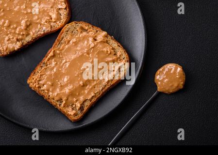 Nahrhaftes Sandwich bestehend aus Brot und Erdnussbutter auf einer schwarzen Keramikplatte auf dunklem Betonhintergrund Stockfoto