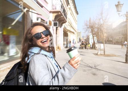 Zufriedene junge Frau mit Rucksack, die eine Kaffeetasse in der Hand hielt, während sie durch die Stadt spazierte. Stockfoto