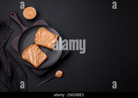 Nahrhaftes Sandwich bestehend aus Brot und Erdnussbutter auf einer schwarzen Keramikplatte auf dunklem Betonhintergrund Stockfoto