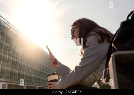 Eine junge Frau mit Sonnenbrille, die auf der Bank sitzt und ihr Handy online nutzt. Stockfoto