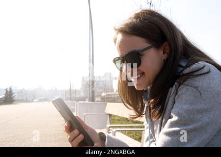 Eine junge Frau mit Sonnenbrille, die auf der Bank sitzt und ihr Handy online nutzt. Stockfoto