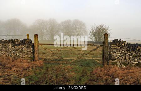 Im nebligen Morgenwinterlicht in den Bergen von Derbyshire schließt ein 5-Bar-Tor einen Feldeingang zwischen trockenen Steinmauern Stockfoto