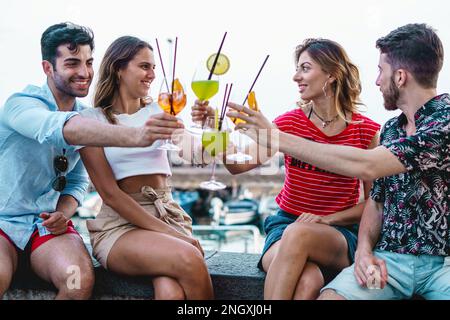 Zwei kaukasische Paare genießen den Sommer auf einer Mauer in der Nähe des Hafens, trinken farbenfrohe Cocktails und lächeln in ihren T-Shirts und Shorts Stockfoto