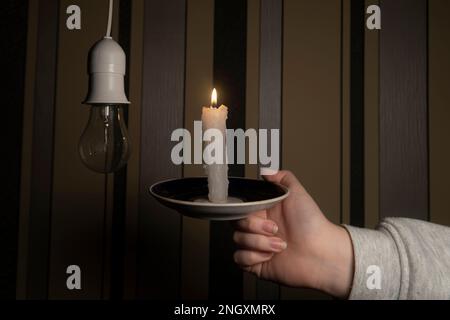 Eine Hand mit brennender Kerze neben einer Patrone mit elektrischer Glühbirne in einem dunklen Raum (Nahaufnahme). Stromausfall. Stromausfallkonzept Stockfoto