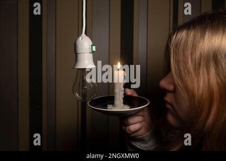 Ein Mädchen in einem dunklen Raum hält eine brennende Kerze in der Hand und schaut mit einer Glühbirne auf den Halter (Nahaufnahme). Stromausfall. Stromausfallkonzept Stockfoto