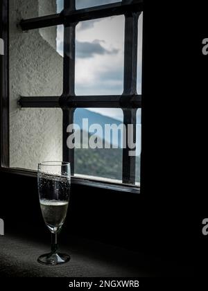 Ein Glas Champagner in einem alten, verschließbaren Burgfenster mit Blick auf die Berge, den bewaldeten Himmel Stockfoto