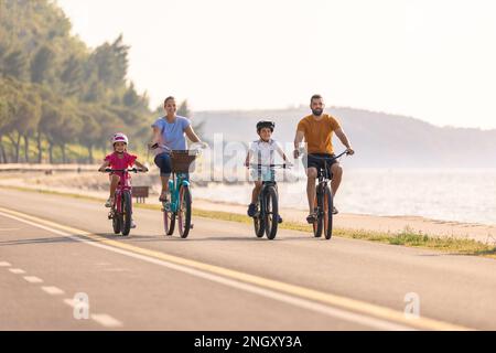 Zwei Kinder mit Helmen auf dem Kopf und lächelnde Eltern, die auf einer familienfreundlichen Radroute entlang der Küste mit Blick auf die Küste Fahrräder fahren. Stockfoto