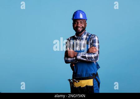 afroamerikanischer Bauarbeiter mit fröhlichem Gesicht, der einen Helm trägt und lächelt, während er in die Kamera schaut. Selbstbewusster Aufbaumeister, der mit einem Schraubenschlüssel mit gekreuzten Armen vor blauem Hintergrund posiert. Stockfoto