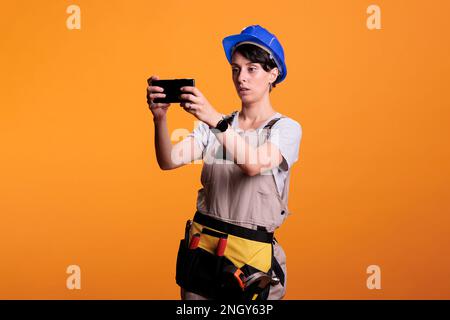 Bauarbeiter, der Fotos und Fotos mit dem Mobiltelefon macht, trägt als Industriehersteller im Studio Schutzhelm. Frau, die als Konstrukteur verkleidet ist und Bilder auf dem Smartphone aufnimmt, fotografiert. Stockfoto