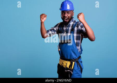 Porträt eines wütenden afroamerikanischen Bauarbeiters, der schreit und seine Hände auf blauem Hintergrund hebt. Builder hat einen Nervenzusammenbruch, frustrierter Mann mit aggressiver Reaktion. Stockfoto