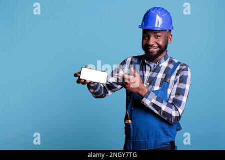 afroamerikanischer Bauarbeiter im Schutzhelm, der mit dem Finger auf das Handy zeigt, mit leerem Bildschirm isoliert auf blauem Hintergrund. Baumeister im Studio mit leerem Werbespot. Stockfoto