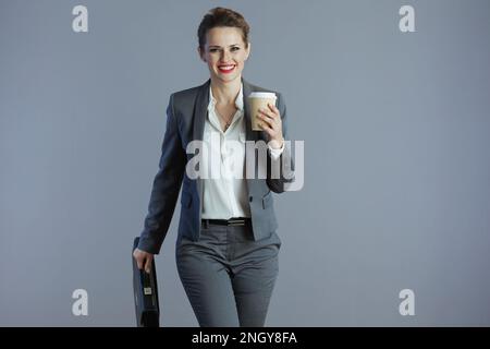 Lächelnde, stilvolle Eigentümerin eines kleinen Unternehmens im grauen Anzug mit Kaffeetasse und Aktentasche vor grauem Hintergrund. Stockfoto