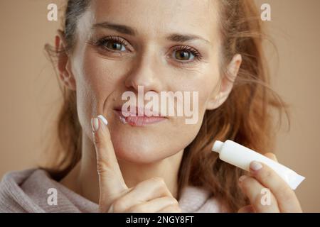 40-jährige Frau mit Herpes auf Lippen und Salbe in einem Schlauch, die Salbe mit dem Finger auf beigem Hintergrund aufträgt. Stockfoto